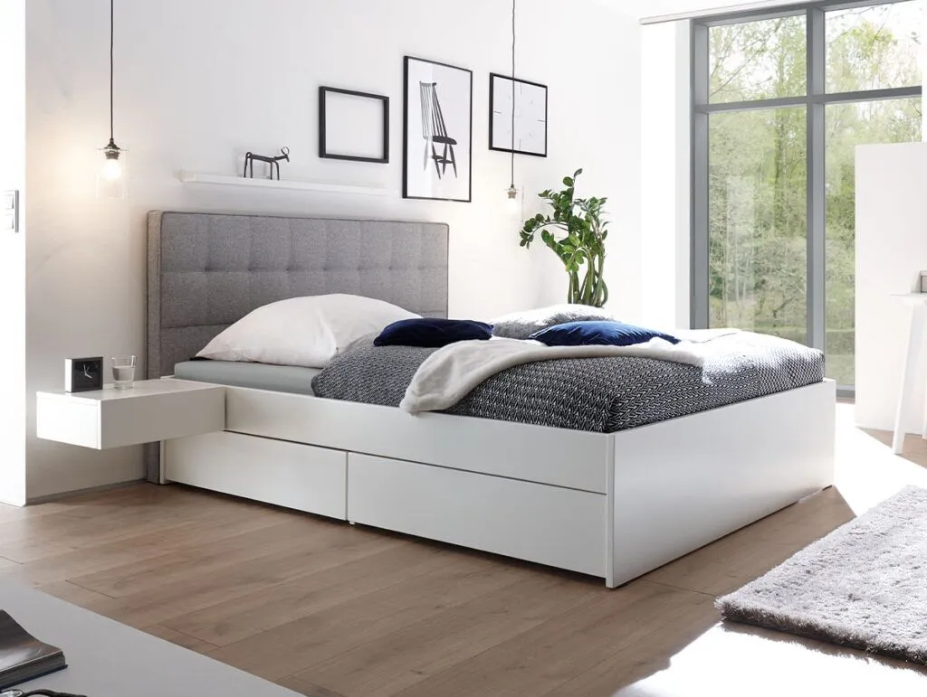 Modernes Schlafzimmer mit dem Hasena Schubladenbett "Function and Comfort Elito 2" mit Schubladen, einem grau gepolsterten Kopfteil, Pendelleuchten, minimalistischem Dekor, großen Fenstern für natürliches Licht und einem Hauch von Grün durch eine Pflanze.