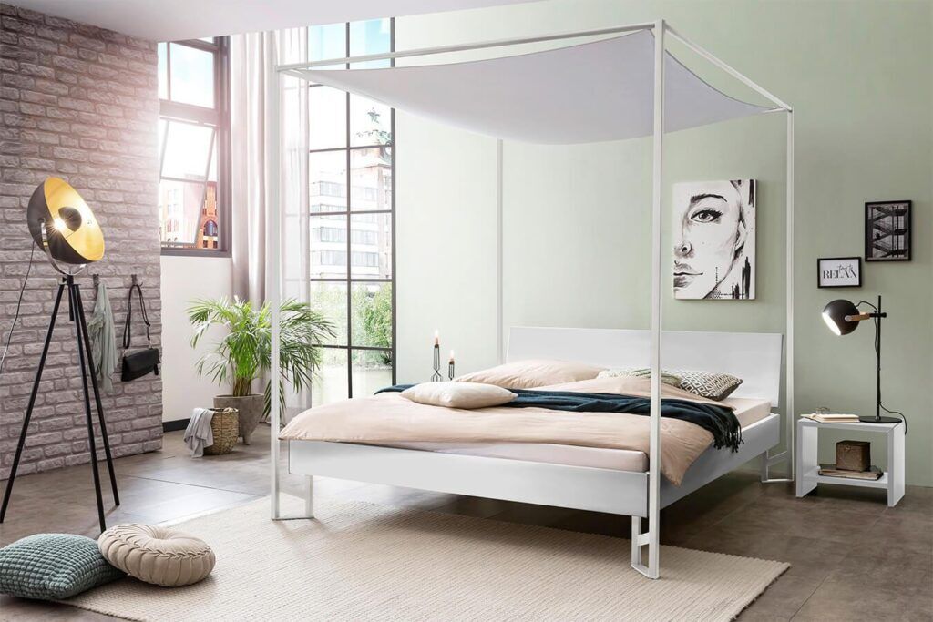 Modernes Schlafzimmer mit weißem Himmelbett, beiger Bettwäsche und einer dunkelgrünen Decke. Große Fenster lassen viel natürliches Licht herein. Zur Ausstattung gehören eine Stehlampe, Zimmerpflanzen, ein Schwarz-Weiß-Porträt und minimalistische Möbel.