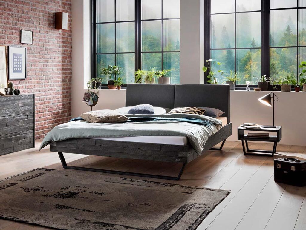 Ein modernes Schlafzimmer mit großen Fenstern, einem Doppelbett mit Kissen und Decken, Topfpflanzen auf der Fensterbank, einem Teppich auf hellem Holzboden, einer Stehlampe und freiliegenden Wänden aus Ziegeln und Holzakzenten.