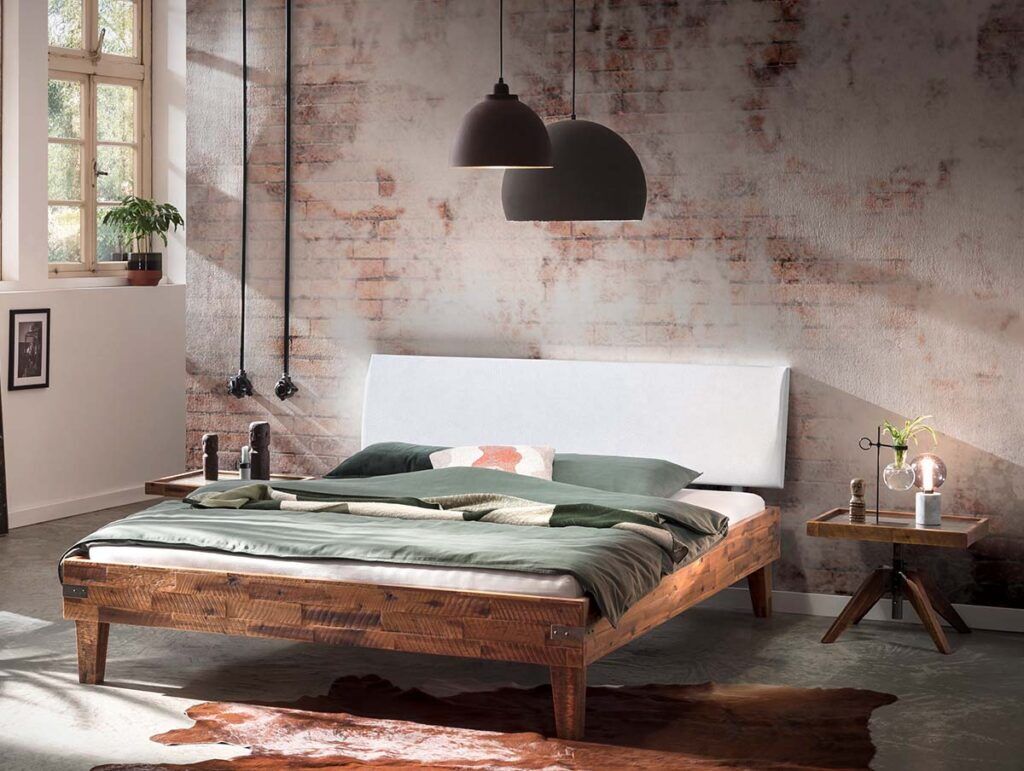 Ein modernes Schlafzimmer mit einem rustikalen Holzbett, grüner und weißer Bettwäsche, zwei Pendelleuchten, einem kleinen Beistelltisch mit Pflanzen und einem Kuhfellteppich auf einem Betonboden. Ein großes Fenster lässt natürliches Licht herein.