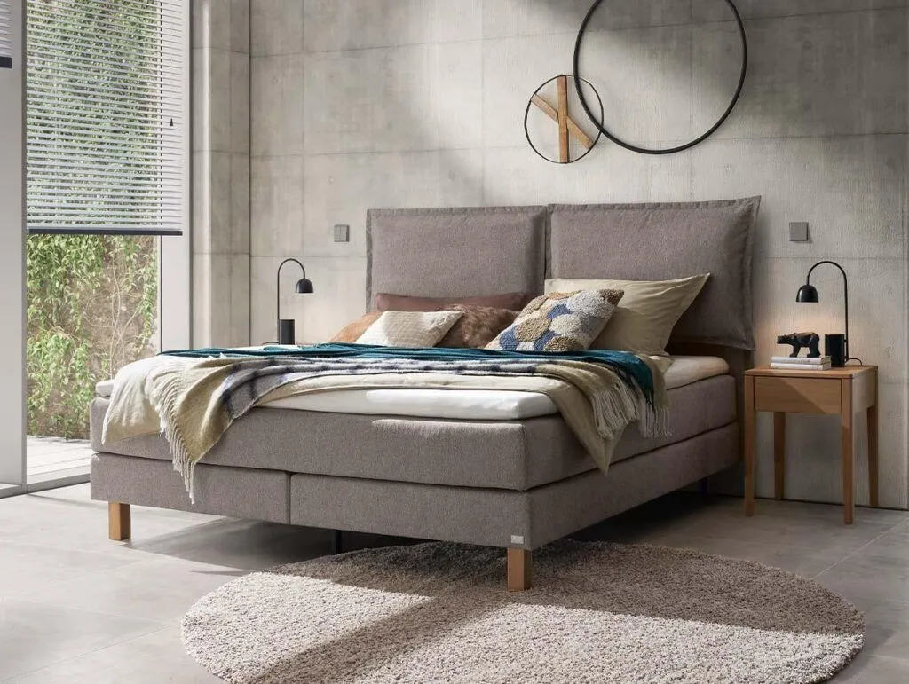 Ein modernes Schlafzimmer mit einem beigen Polsterbett, verschiedenen Kissen und einer blauen Überwurfdecke. Es gibt zwei hölzerne Nachttische mit schwarzen Lampen, einen runden Spiegel an der Wand und einen runden beigen Teppich auf einem hellen Fliesenboden.