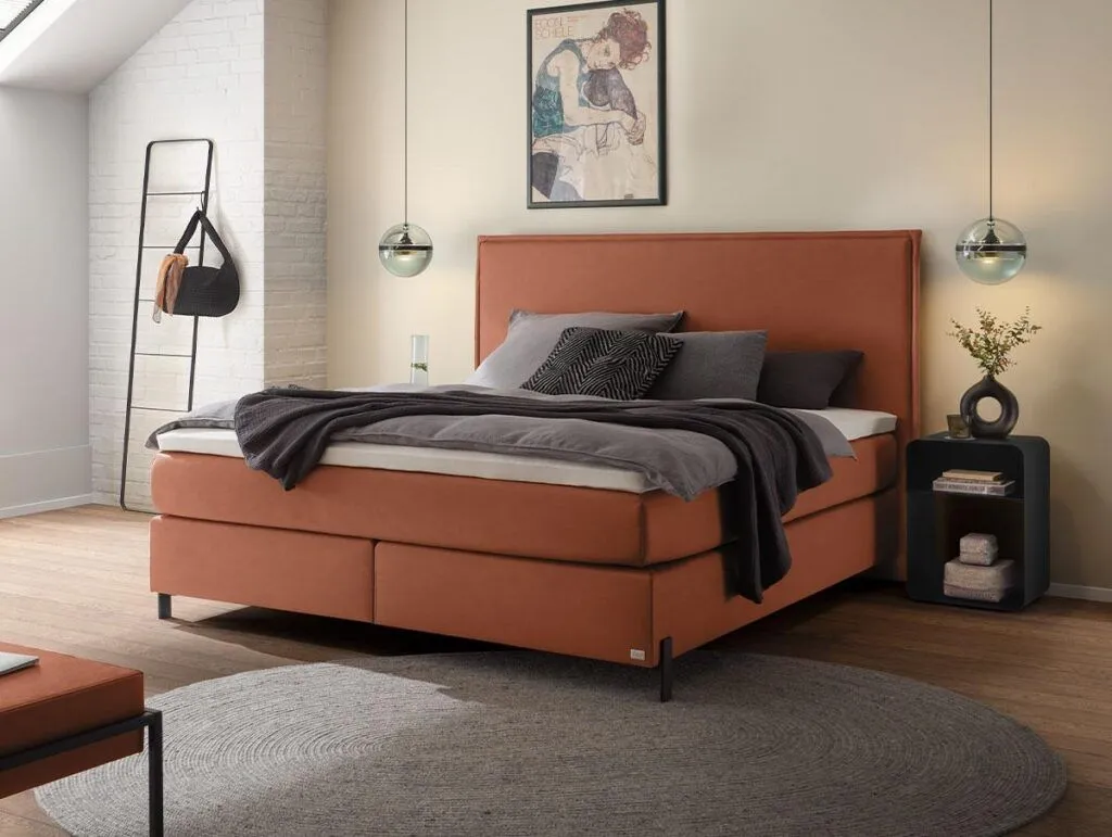 Ein modernes Schlafzimmer von Bettenland verfügt über ein rostfarbenes Bett, graue Bettwäsche, Pendelleuchten, einen runden grauen Teppich, einen kleinen schwarzen Nachttisch mit einer Vase und ein gerahmtes Kunstwerk an der beigen Wand über dem Bett.