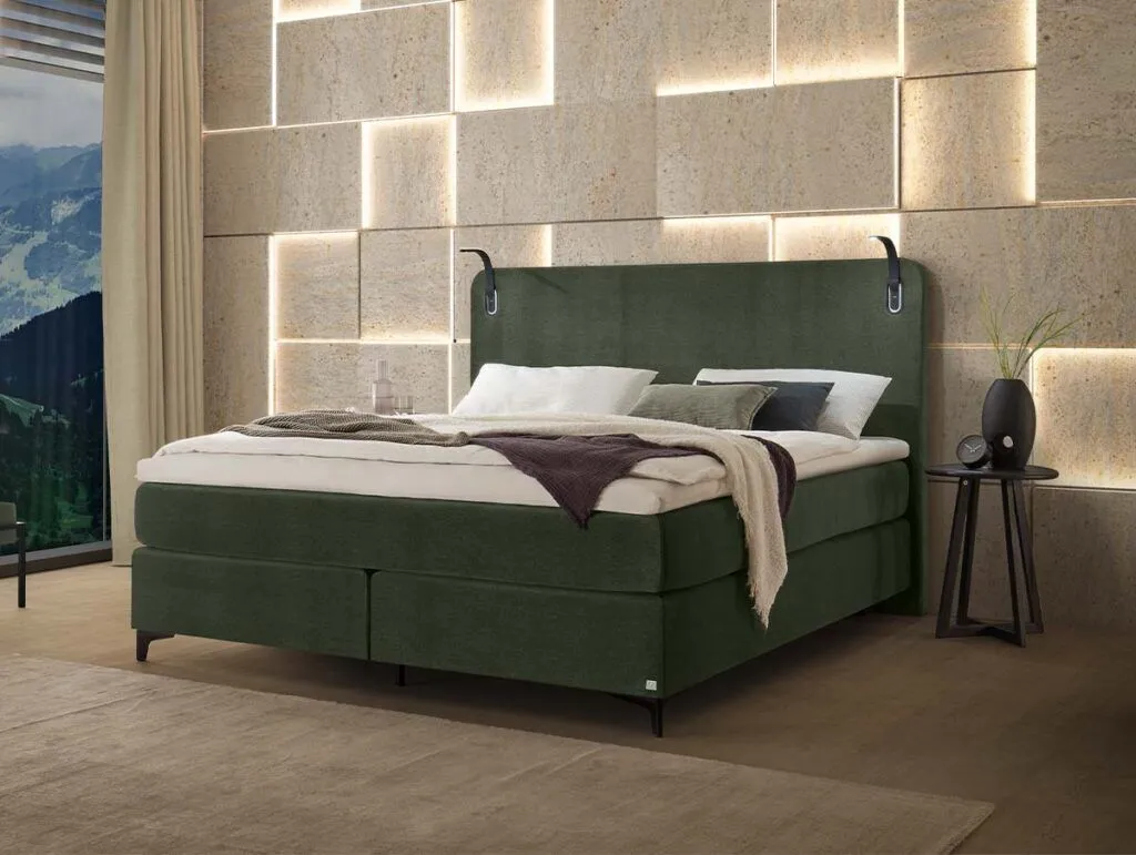 Ein modernes Schlafzimmer mit einem grün gepolsterten Bett, das mit weißer und grauer Bettwäsche ausgelegt ist. An der Wand dahinter befinden sich geometrische Steinplatten mit warmer Hintergrundbeleuchtung. Auf einem runden schwarzen Beistelltisch stehen eine Vase und ein Glas, und ein Fenster gibt den Blick auf die Landschaft frei.