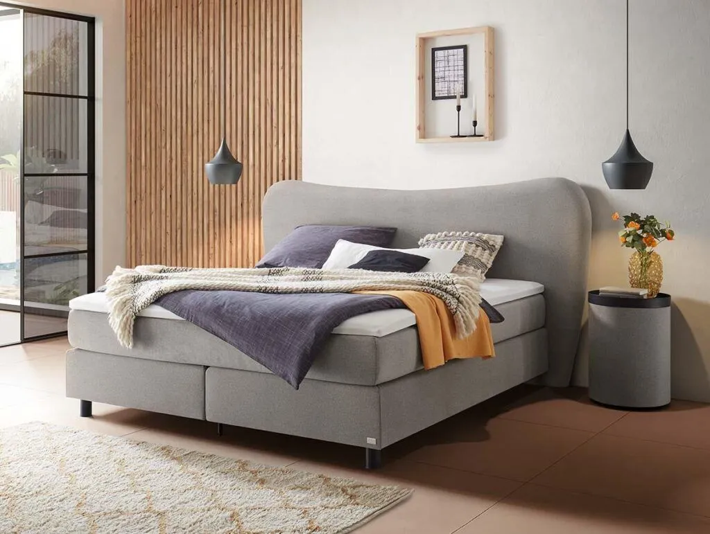 Ein modernes Schlafzimmer mit einem großen grauen Bett, mehreren Kissen und Decken, zwei schwarzen Pendelleuchten, einem runden Nachttisch mit einer Blumenvase und einem Teppich auf einem hellen Boden. Hölzerne und weiße Wände sorgen für eine gemütliche Atmosphäre.