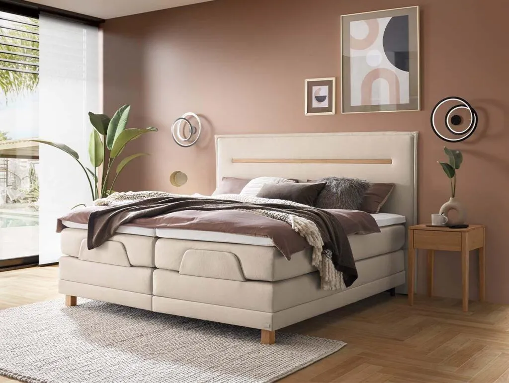 Ein modernes Schlafzimmer mit einem beigen Polsterbett, brauner und grauer Bettwäsche, einem hölzernen Nachttisch, Wandkunst, grünen Pflanzen und runden Wandleuchten vor braunen Wänden und großen Fenstern mit natürlichem Licht.