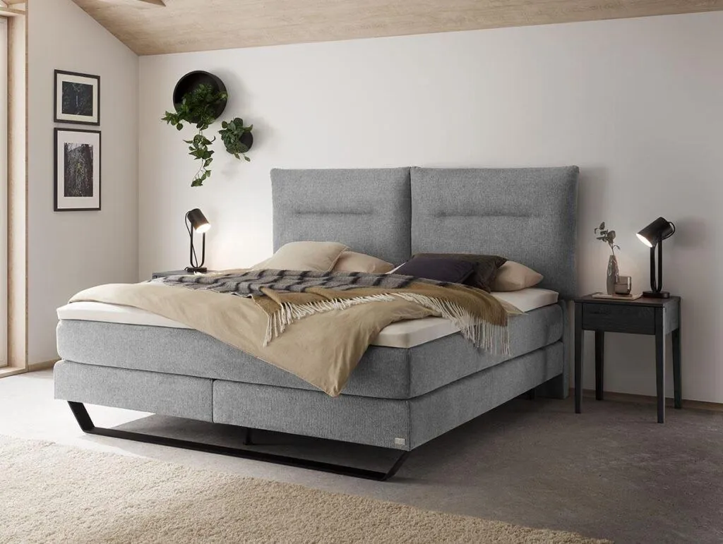 Ein modernes Schlafzimmer mit einem grauen Polsterbett, beigen Decken, zwei schwarzen Nachttischen mit Lampen, einer Wandpflanze und gerahmter Kunst an einer weißen Wand. Das Zimmer hat eine minimalistische, gemütliche Atmosphäre.