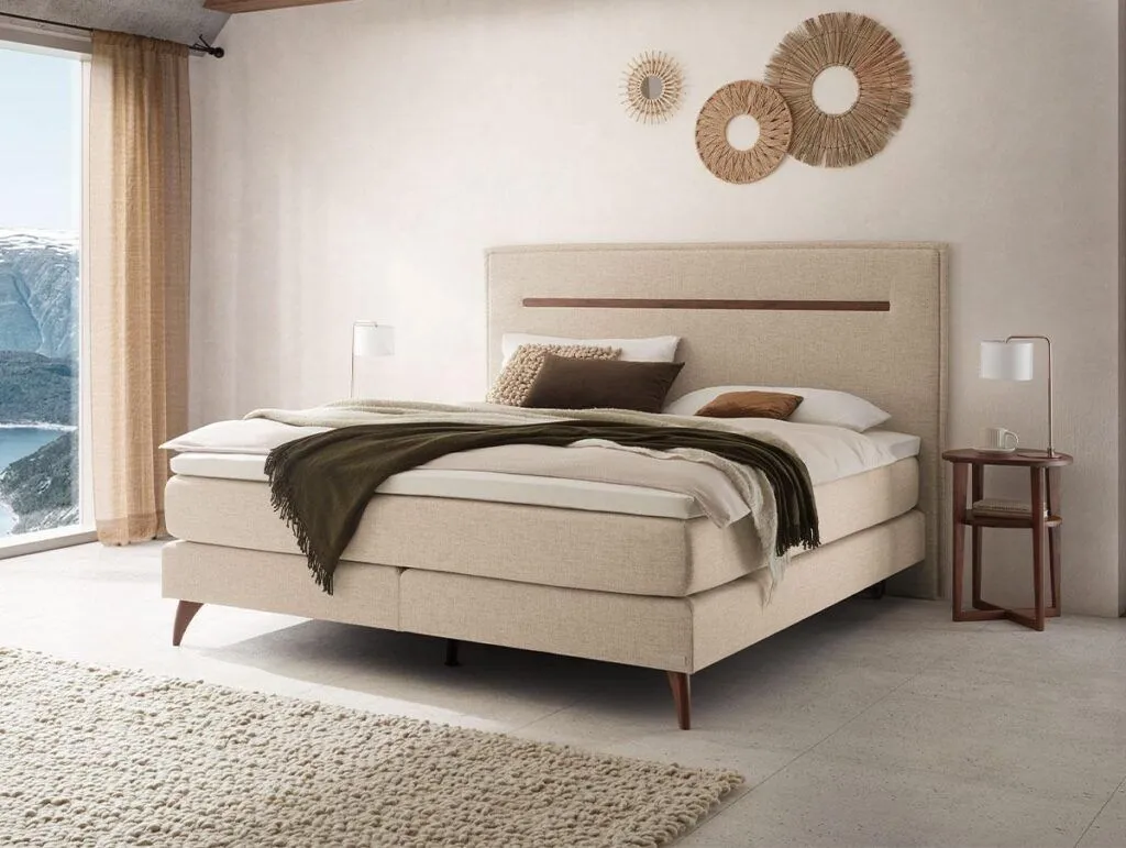 Ein modernes beiges Bett mit weißer und brauner Bettwäsche steht in einem hellen, minimalistischen Schlafzimmer. Dekorative Wandkunst hängt über dem Bett, und das Sonnenlicht strömt durch große Fenster neben transparenten Vorhängen.