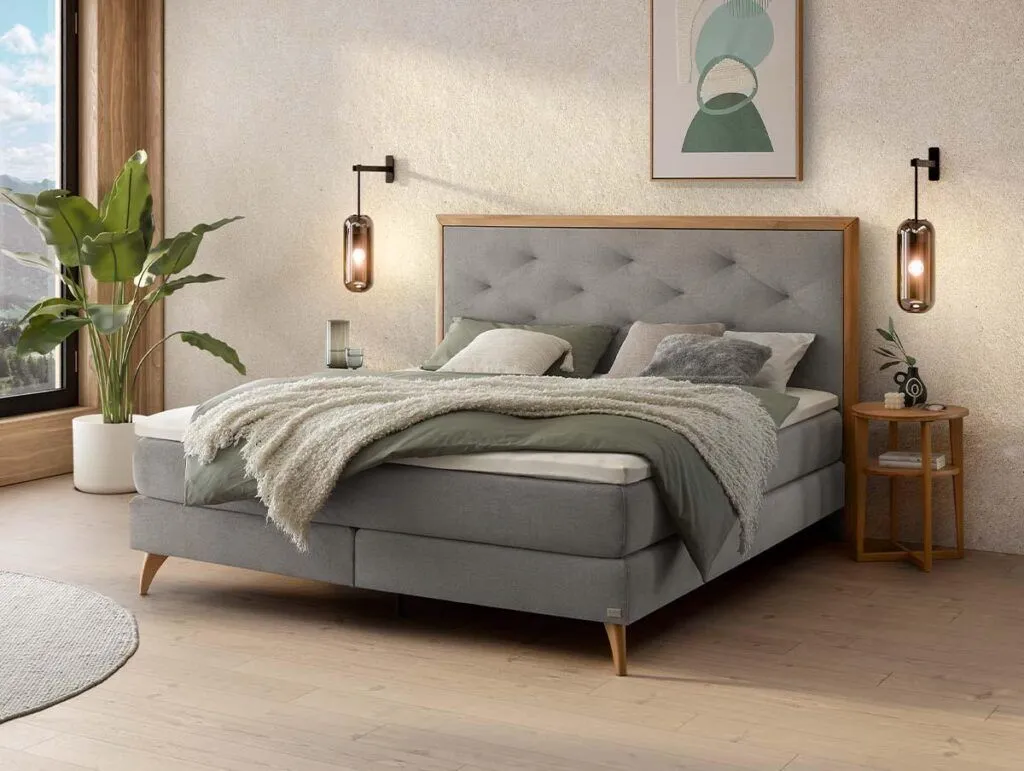 Ein modernes Schlafzimmer mit einem grauen Polsterbett, grüner und beiger Bettwäsche, Nachttischen, Hängelampen, einer Topfpflanze und abstrakter Wandkunst. Natürliches Licht fällt durch ein großes Fenster ein.