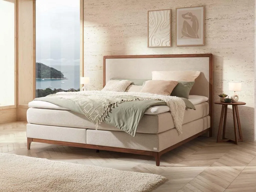 Ein modernes Schlafzimmer mit einem großen gepolsterten Bett, neutraler Bettwäsche und sanften grünen Akzenten. Es gibt zwei Nachttische mit Lampen, abstrakte Wandkunst und große Fenster mit Blick auf das Meer und die Hügel.