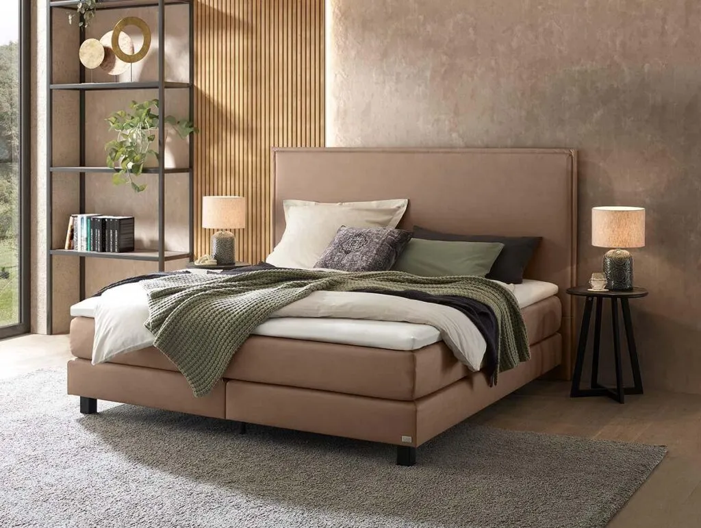 Ein modernes Schlafzimmer mit einem beigen Polsterbett, das mit Kissen und einer grünen Strickdecke ausgelegt ist. Zwei runde Nachttische mit Lampen flankieren das Bett. Ein Regal mit Pflanzen und Büchern steht neben einem großen Fenster.