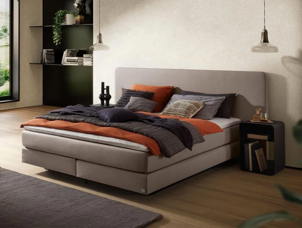 Ein modernes Schlafzimmer mit einem beigen Polsterbett mit orangefarbener, grauer und schwarzer Bettwäsche, zwei Hängelampen, einem kleinen schwarzen Nachttisch und einem Regal mit Büchern und Pflanzen vor einer hellen Wand.