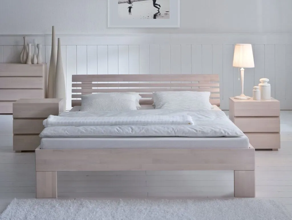 Ein minimalistisches Schlafzimmer mit dem Hasena Bett "Wood-Line Classic Massa" Holzbettgestell mit weißer Bettwäsche, zwei Nachttischen, einer Lampe, Vasen, einer Kommode und monochromer Wandkunst für einen klaren, hellen und modernen Look mit hellem Dekor.