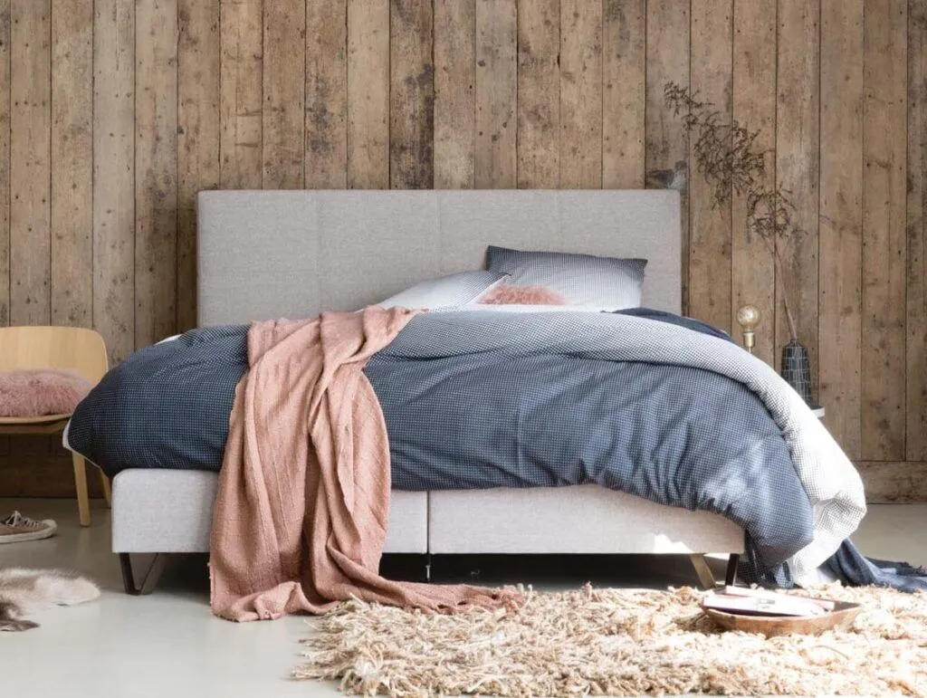 Ein gemütliches Schlafzimmer mit einem grauen Polsterbett, blauer Bettwäsche und einer rosa Überwurfdecke. Das Zimmer verfügt über eine Wand aus Holzdielen, einen beigen Teppich, einen einfachen Stuhl mit einem rosa Kissen und einen kleinen Nachttisch mit Dekoration.
