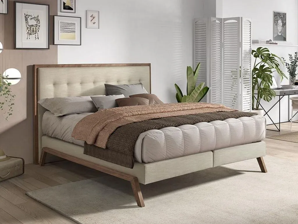 Ein modernes Schlafzimmer mit einem beigen Polsterbett, neutraler Bettwäsche und einer braunen Decke. Das Zimmer hat gerahmte Kunstwerke an der Wand, Pflanzen, einen Raumteiler und einen Schreibtisch in der Ecke, alles auf hellem Holzboden mit einem beigen Teppich.