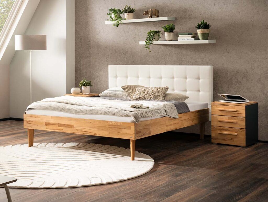 Ein modernes Schlafzimmer mit einem Holzbett, einem weißen getufteten Kopfteil, gemütlicher Bettwäsche, einem runden weißen Teppich, einem hölzernen Nachttisch, zwei Wandregalen mit Pflanzen, einer Lampe und Sonnenlicht, das durch ein großes Fenster fällt.