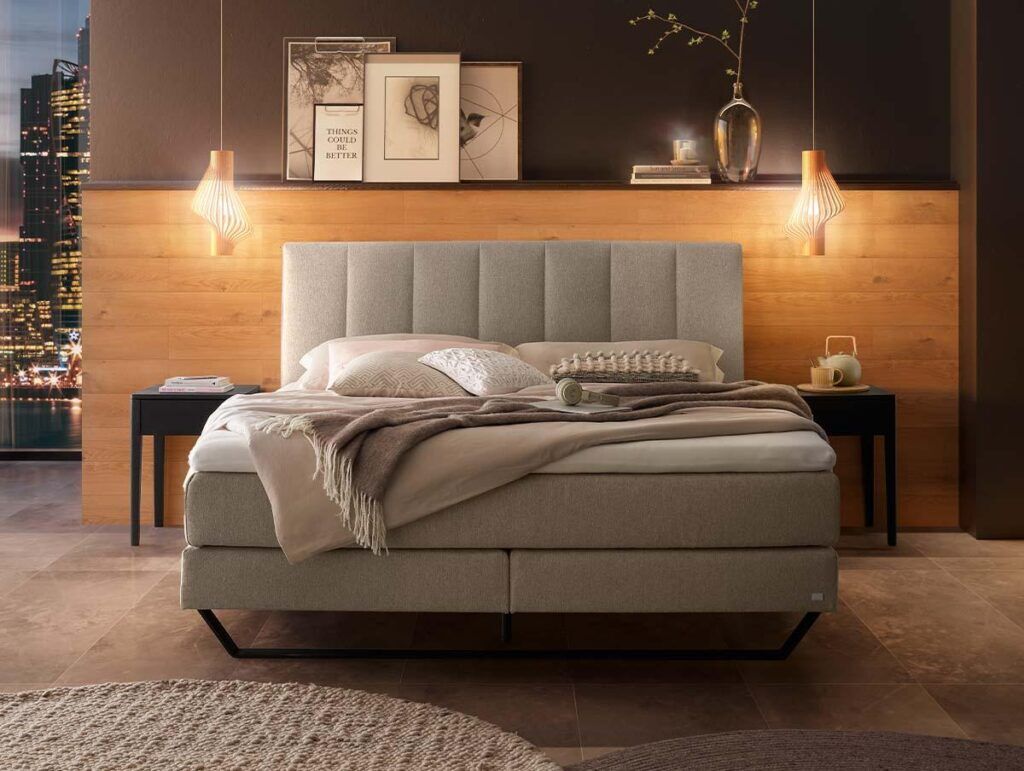 Ein modernes Schlafzimmer mit einem beigefarbenen Polsterbett, kuscheligen Kissen und Decken, zwei Pendelleuchten, Nachttischen mit Tassen und Dekoration, einer holzgetäfelten Wand, gerahmter Kunst und einer durch ein Fenster sichtbaren Stadtlandschaft.