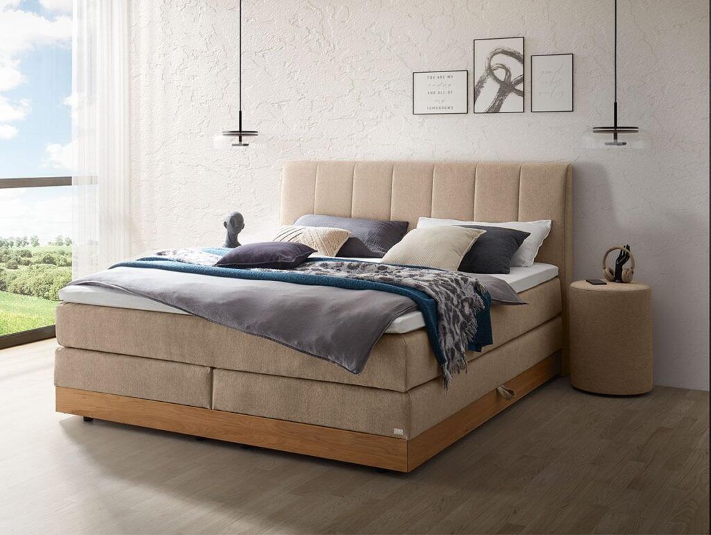 Ein modernes Schlafzimmer mit einem beigen Polsterbett, ordentlich angeordneten Kissen, einer blau-grauen Decke, einem runden beigen Nachttisch, zwei Pendelleuchten und einem großen Fenster mit Blick ins Freie.