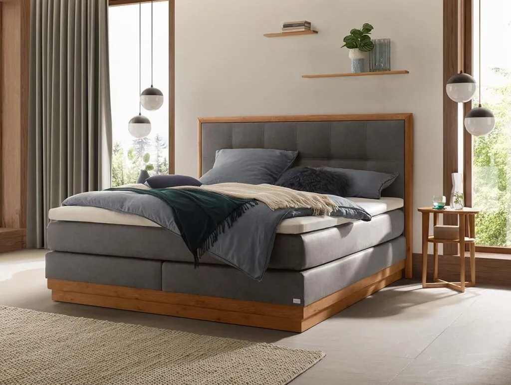 Ein modernes Schlafzimmer mit dem Hasena Boxspringbett Modern "Gavin Leona", das ein grau gepolstertes Kopfteil und einen Holzrahmen aufweist. Zwei Pendelleuchten flankieren das Bett neben einem kleinen Beistelltisch aus Holz und großen Fenstern mit Blick nach draußen.
