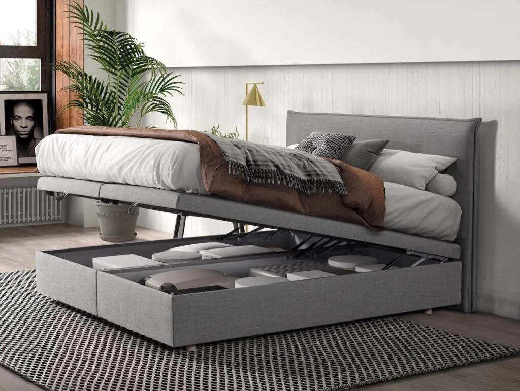 Ein graues gepolstertes Bett, dessen Matratze hochgeklappt ist, so dass darunter ein Stauraum mit Kisten zum Vorschein kommt. Im Zimmer stehen eine Pflanze, ein gerahmtes Foto, ein Fenster mit Sonnenlicht und eine neutrale Einrichtung.