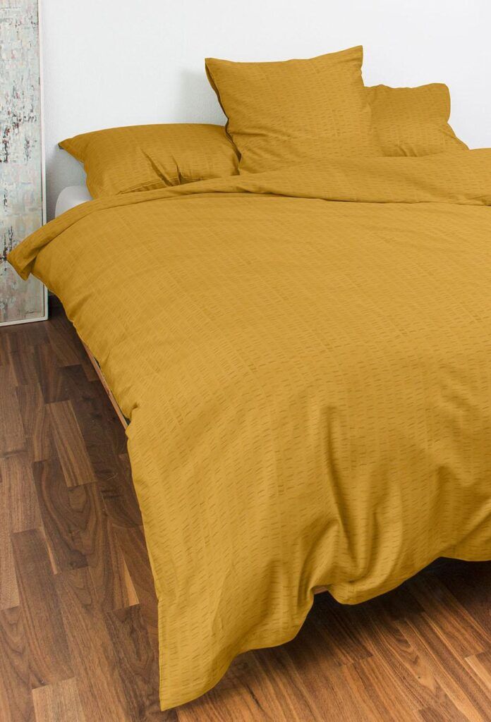 Ein ordentlich gemachtes Bett mit Bettwäsche Night Fashion Edi in Senfgelb, mit Bettdecke und Kissen, steht auf einem Holzboden neben einer weißen Wand und einem abstrakten Kunstwerk.