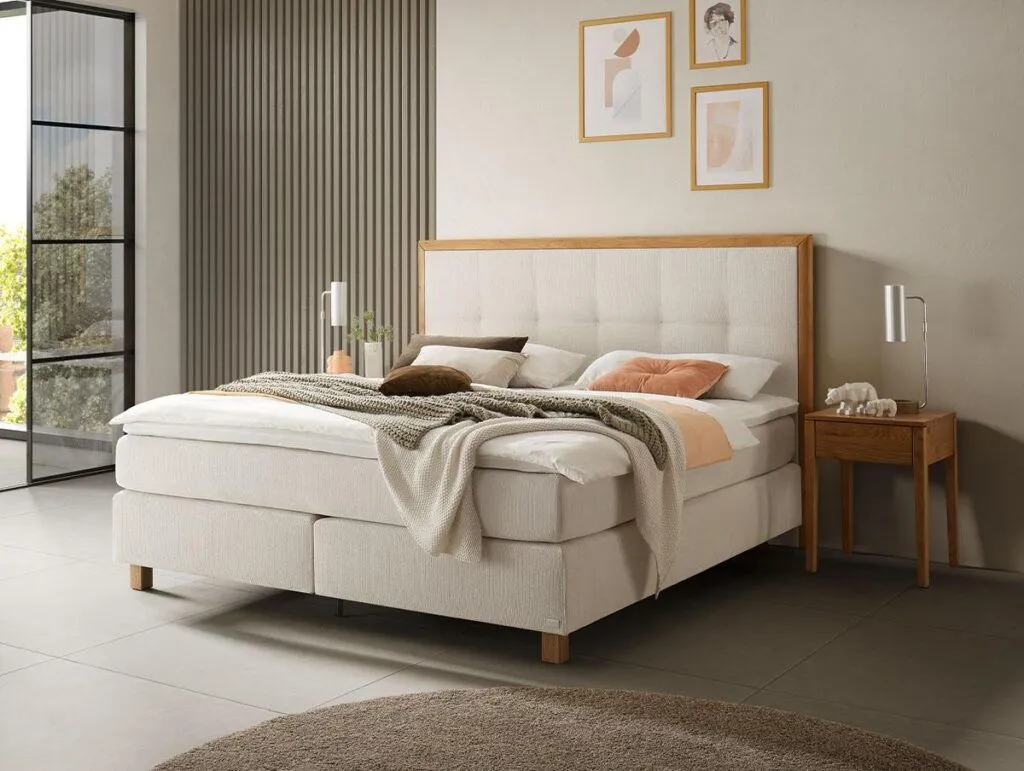 Ein modernes Schlafzimmer mit einem großen Polsterbett, beiger Bettwäsche, einem hölzernen Nachttisch mit einer Lampe, gerahmter Kunst an der Wand und einem runden braunen Teppich auf einem gefliesten Boden. Das Sonnenlicht scheint durch ein großes Fenster.