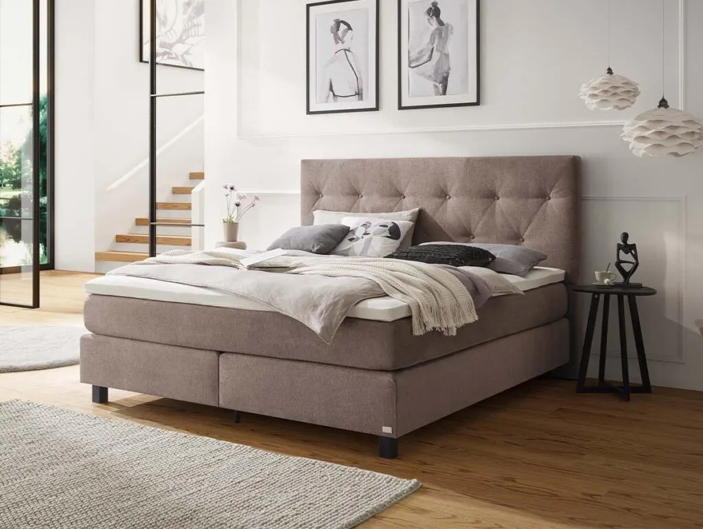 Ein modernes, gemütliches Schlafzimmer mit einem taupefarbenen gepolsterten Bett, das mit neutraler Bettwäsche und Kissen ausgestattet ist. Über dem Bett hängen drei schwarz-weiße Wandgemälde. Durch die großen Fenster fällt Licht herein und erhellt den Holzboden.