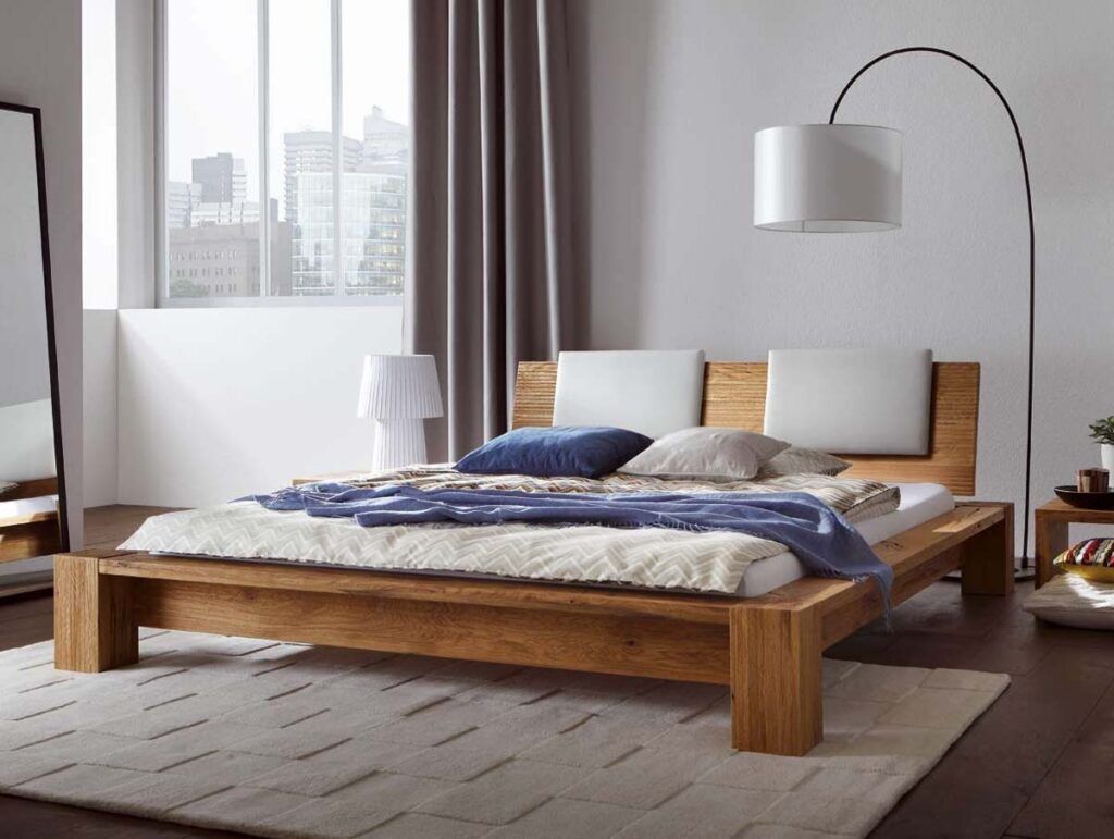 Ein modernes Schlafzimmer mit dem Hasena Bett "Oak-Wild Cortina 2", einem niedrigen Holzbett, weißer und blauer Bettwäsche, einer Stehlampe, einem großen Fenster mit Blick auf die Stadt, grauen Vorhängen, einem weichen beigen Teppich, einem Spiegel und einer weißen Lampe am Fenster.