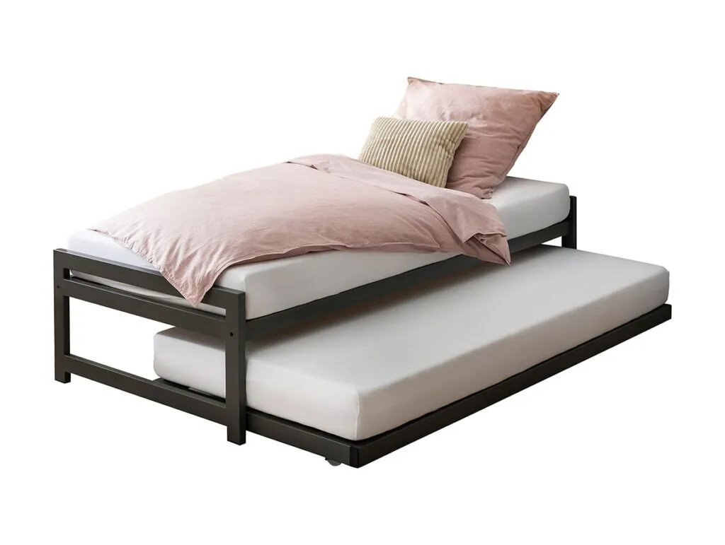 Ein schwarzes Metallbett mit einer rosa Bettdecke, einem Kissen und einem beigen Kissen auf der oberen Matratze; die untere Matratze ist schlicht weiß und wird von unten herausgezogen.