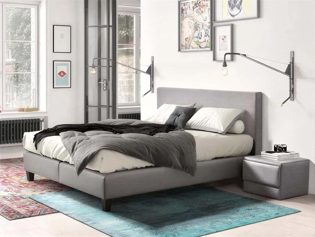 Modernes Schlafzimmer mit grauem Polsterbett, bunten Teppichen, gerahmter Kunst an der Wand, großen Fenstern mit natürlichem Licht, Pflanzen in Regalen und einer gemütlichen, einladenden Atmosphäre. Ein Karton und eine Einkaufstasche stehen auf dem Boden.