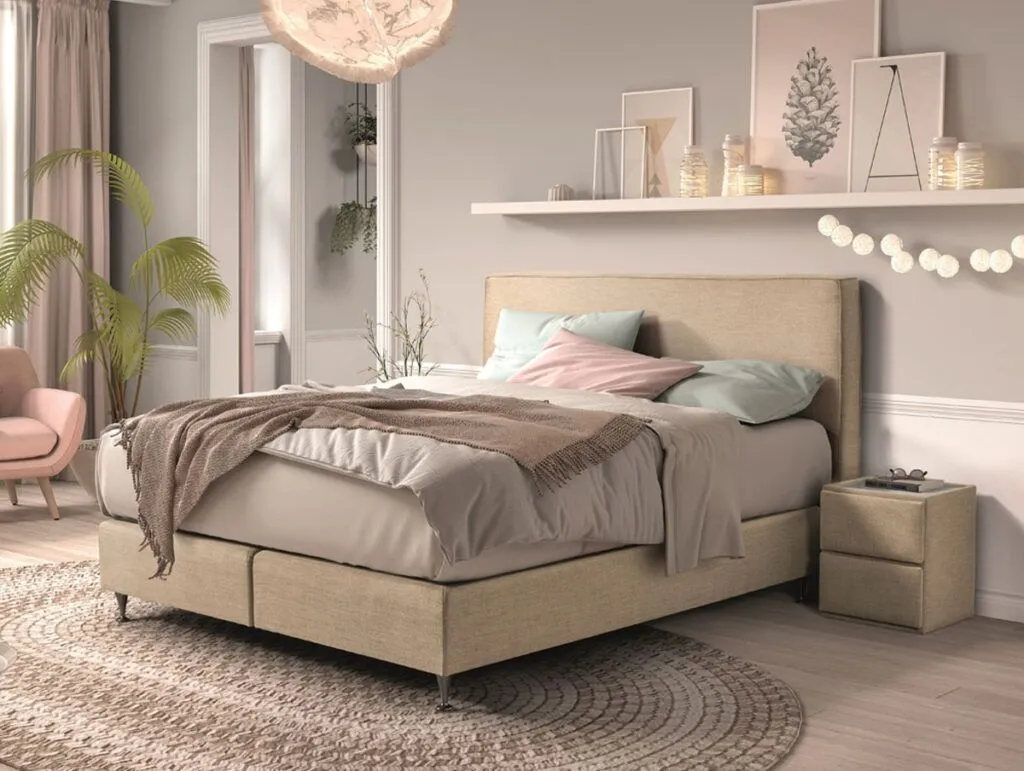 Ein gemütliches, modernes Schlafzimmer mit einem beigen Bett, weicher Bettwäsche und einer taupefarbenen Überwurfdecke. Es gibt einen runden gewebten Teppich, einen rosafarbenen Sessel, üppige Pflanzen, Kerzen und ein schwebendes Regal mit Kunstwerken und dekorativen Leuchten über dem Bett.