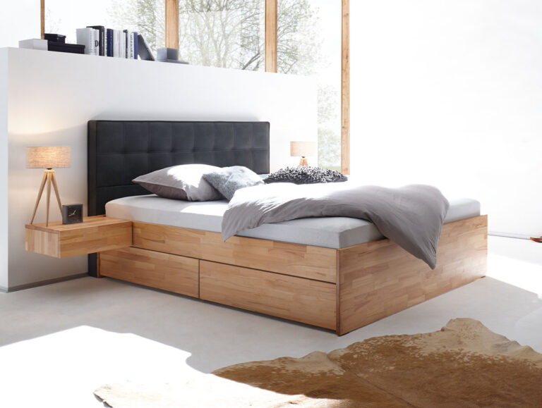 Ein modernes Schlafzimmer mit einem Hasena Schubladenbett "Function and Comfort Elito 2" mit Schubladen, grauem Polsterkopfteil, grauer Bettwäsche, zwei Nachttischen mit Lampen, einem Kuhfellteppich auf hellem Boden und großen Fenstern, die natürliches Licht hereinlassen.