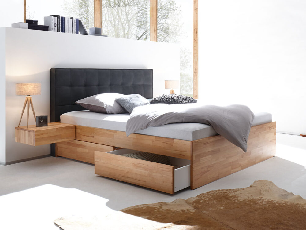 Ein modernes Schlafzimmer mit einem Hasena Schubladenbett "Function and Comfort Elito 2" mit eingebauten Schubladen, grauem Polsterkopfteil, grauer Bettwäsche, schwebendem Nachttisch mit Lampe, Kuhfellteppich und großen Fenstern für natürliches Licht.