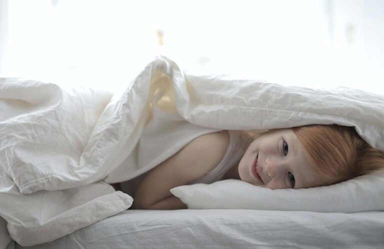 Ein kleines Kind mit roten Haaren liegt im Bett unter einer weißen Decke, lächelt und stützt seinen Kopf auf ein weißes Kissen. Das Zimmer ist hell und sanft beleuchtet - einfache Tipps für gesunden und erholsamen Schlaf umgesetzt.