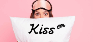 Eine Frau mit einer rosafarbenen Schlafmaske auf dem Kopf hält ein weißes Kissen mit der Aufschrift Kiss em vor einem rosafarbenen Hintergrund - Kisskauf leicht gemacht!.