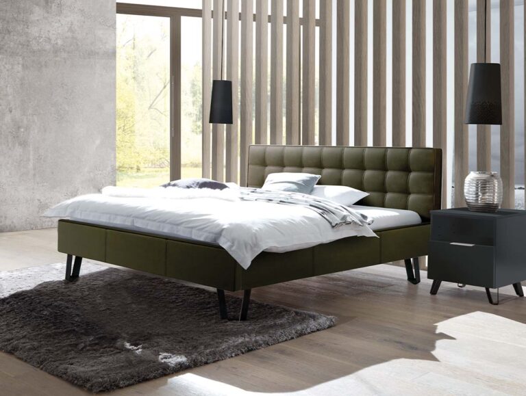 Ein modernes Schlafzimmer mit dem Hasena Bett "Dream-Line Lungo 23 Jeno" mit grüner Polsterung, weißer Bettwäsche, einem grauen Teppich, einem schwarzen Nachttisch mit Vase, zwei schwarzen Stehlampen und großen Fenstern, die natürliches Licht hereinlassen.