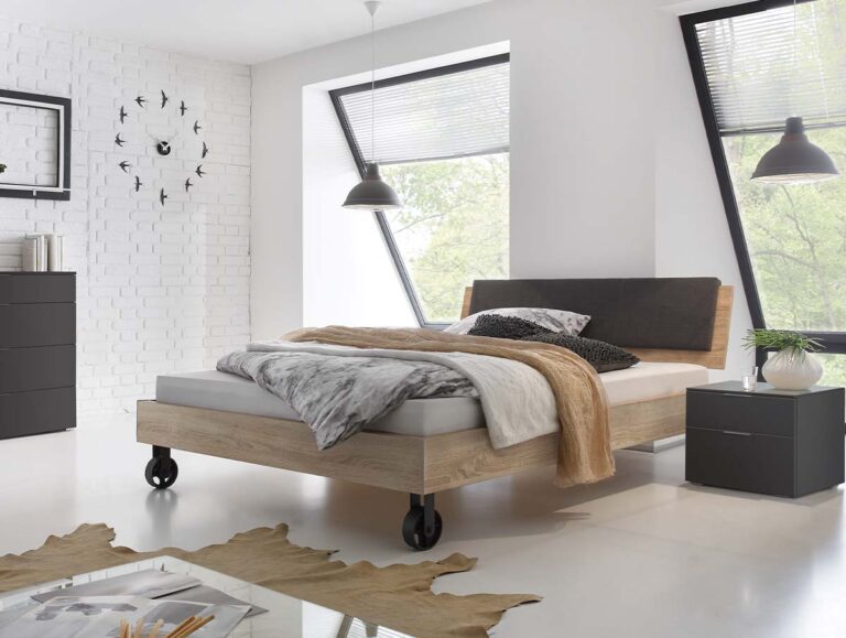 Modernes Schlafzimmer mit großen schrägen Fenstern, einem Hasena Bett "Top-Line Advance Road" mit Holzrahmen und grauem Kopfteil, neutraler Bettwäsche, hellbrauner Decke, Nachttisch, schwarzer Kommode, Wanduhr, Hängeleuchten und einem Kuhfellteppich auf weißem Boden.