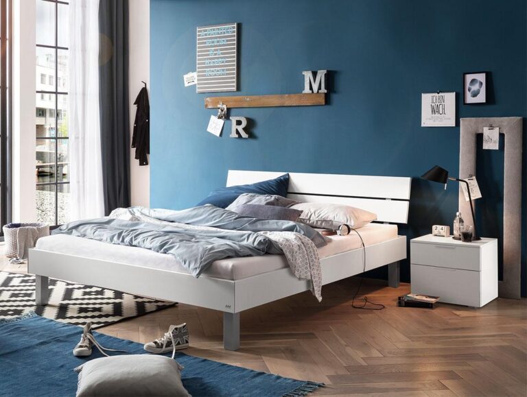 Ein modernes Schlafzimmer mit einem weißen Bett, einer blauen Akzentwand und einem weißen Nachttisch. Das Bett ist mit blauer und grauer Bettwäsche bezogen. Kunst, Buchstaben und Dekoration hängen an der Wand. Sonnenlicht strömt durch hohe Fenster mit transparenten Vorhängen.