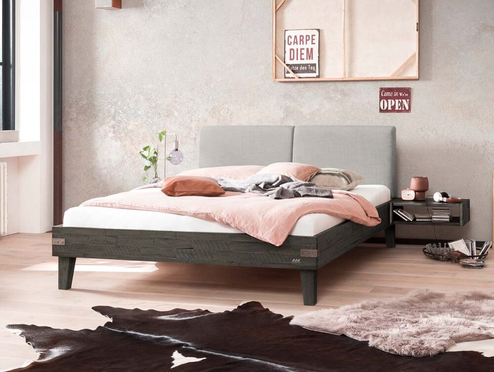 Ein modernes Schlafzimmer mit einem grauen Polsterbett, weißer und roter Bettwäsche und einem hölzernen Nachttisch. An der Wand hängen ein "Carpe Diem"-Schild und strukturierter Beton. Auf dem hellen Holzboden liegen ein Kuhfellteppich und ein rosafarbener Flauschteppich.