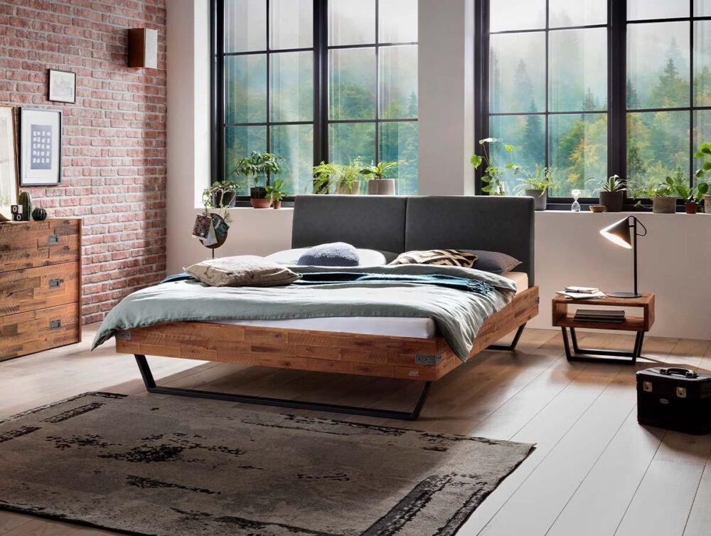 Ein modernes Schlafzimmer mit einem Holzbett, einem grauen Kopfteil und gemütlicher Bettwäsche. Große Fenster lassen natürliches Licht herein und zeigen die Bäume draußen. Topfpflanzen, ein Teppich, ein Nachttisch, eine Lampe und eine Kommode vervollständigen den Raum.