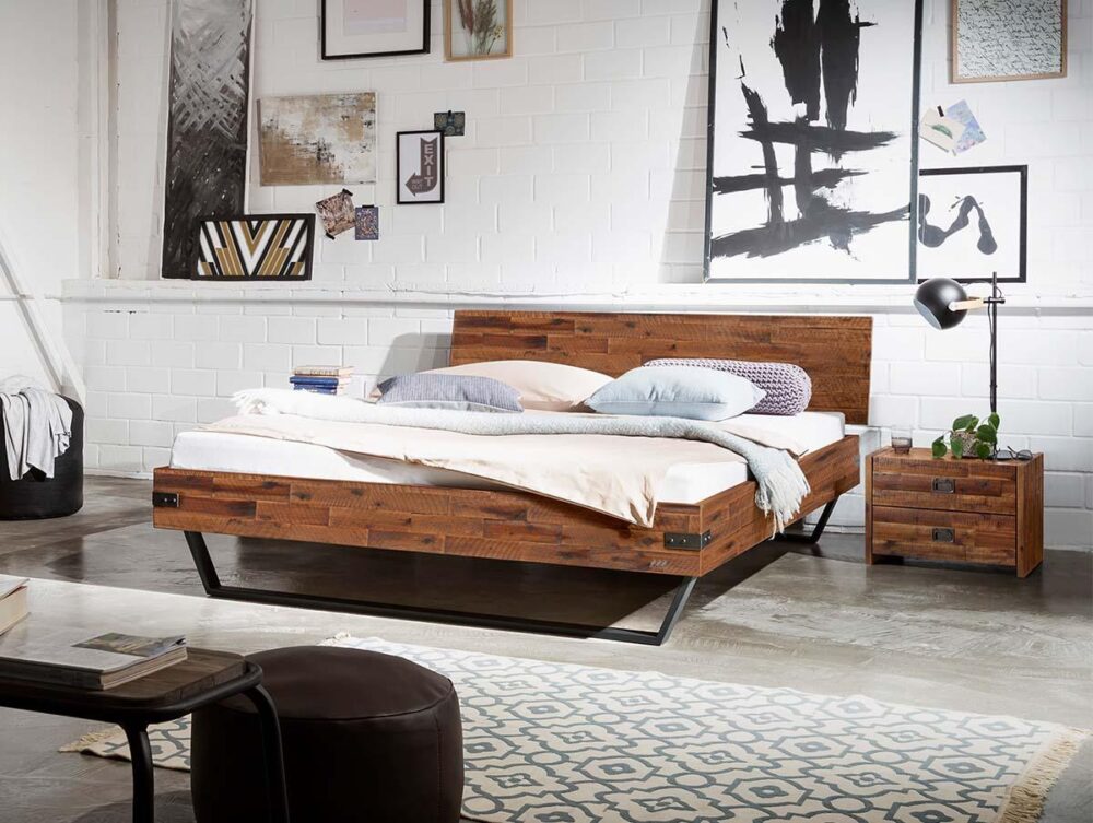 Ein modernes Schlafzimmer mit einem Holzbettgestell, weißer Bettwäsche und Kissen. Das Zimmer hat weiße Backsteinwände, abstrakte Kunst, einen Nachttisch mit einer Lampe und dekorative Gegenstände für einen gemütlichen, modernen Look.
