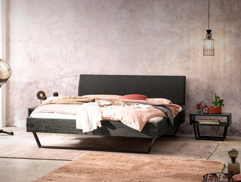 Ein modernes Schlafzimmer mit einem Bettgestell aus dunklem Holz, neutraler Bettwäsche, einer rosa Decke, einem Beistelltisch mit einer Pflanze, einer von der Decke hängenden Lampe und einer strukturierten hellrosa Wand. Das Licht fällt von links ein.