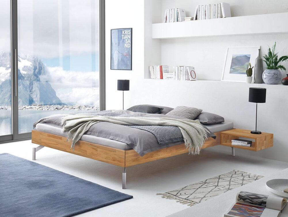 Minimalistisches Schlafzimmer mit einem Holzpritschenbett, grauer Bettwäsche, zwei schwarzen Lampen, Wandregalen mit Büchern, einem großen Fenster, das Berge und Wasser zeigt, und zwei Teppichen auf weißem Boden.