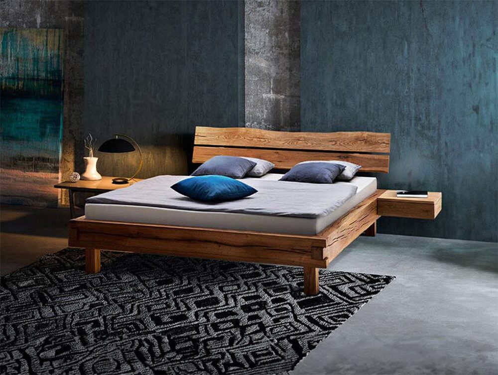 Ein modernes Schlafzimmer mit einem Holzbettgestell, einer grauen Matratze und blauen und grauen Kissen. Das Bett steht auf einem schwarz-weiß gemusterten Teppich neben einem Nachttisch mit einer Lampe, Büchern und einer Vase vor blau-grauen, strukturierten Wänden.