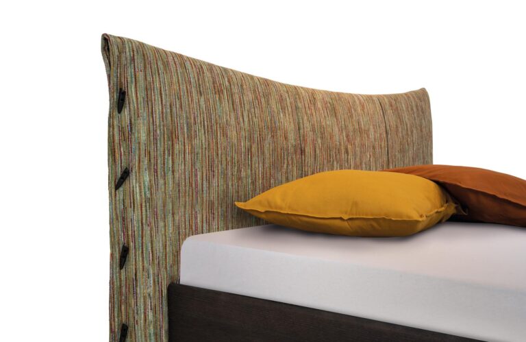 Ein modernes Bett mit einem mehrfarbigen, strukturierten Kopfteil, einer weißen Matratze und zwei Kissen - einem gelben und einem braunen - vor einem weißen Hintergrund.