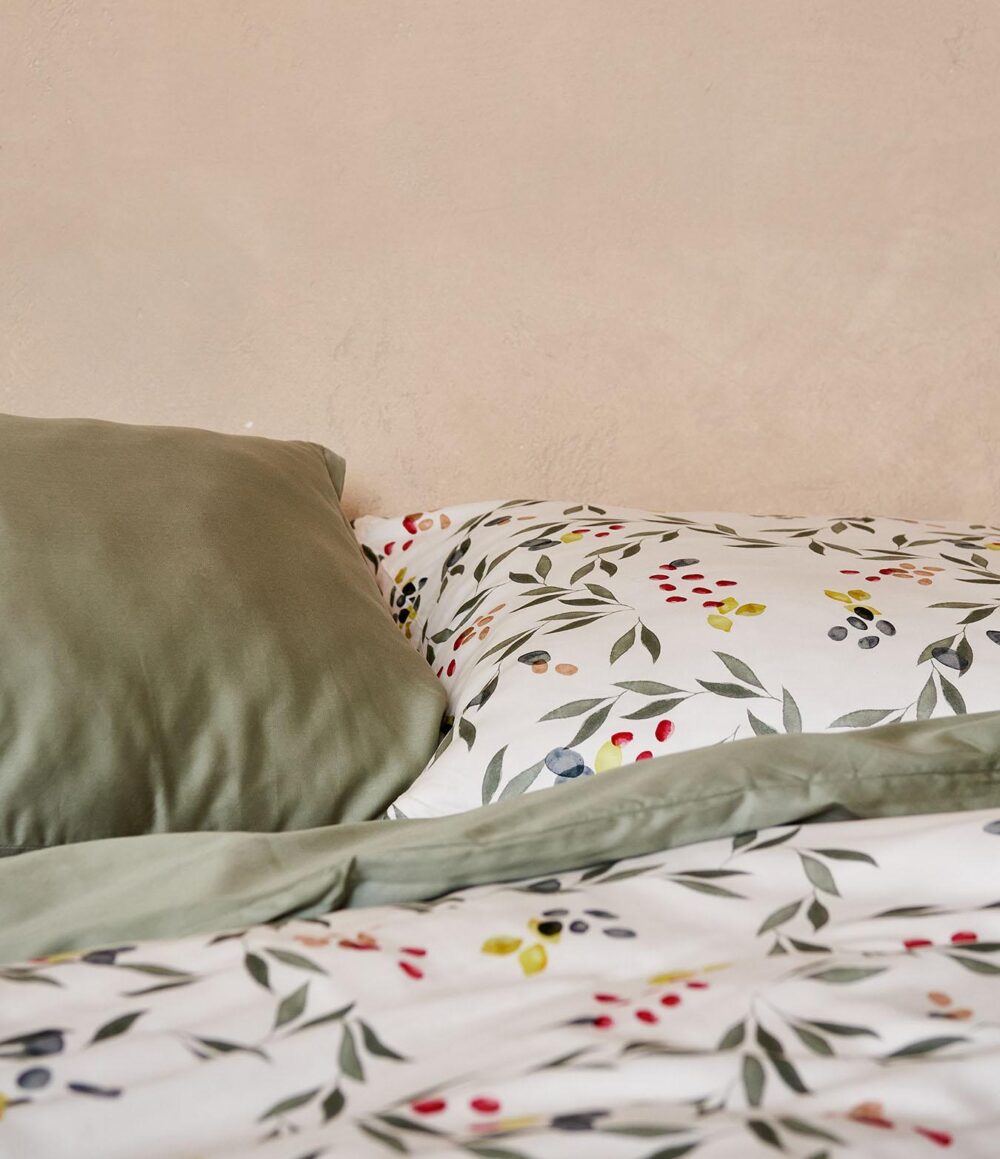 Ein ordentlich gemachtes Bett mit Bettwäsche Journey Living Amiata: ein grüner Kissenbezug und eine Bettdecke, gepaart mit weißen Laken, alles verziert mit einem lebhaften botanischen Blätter- und Beerenmuster, vor einer hellbeigen Wand.