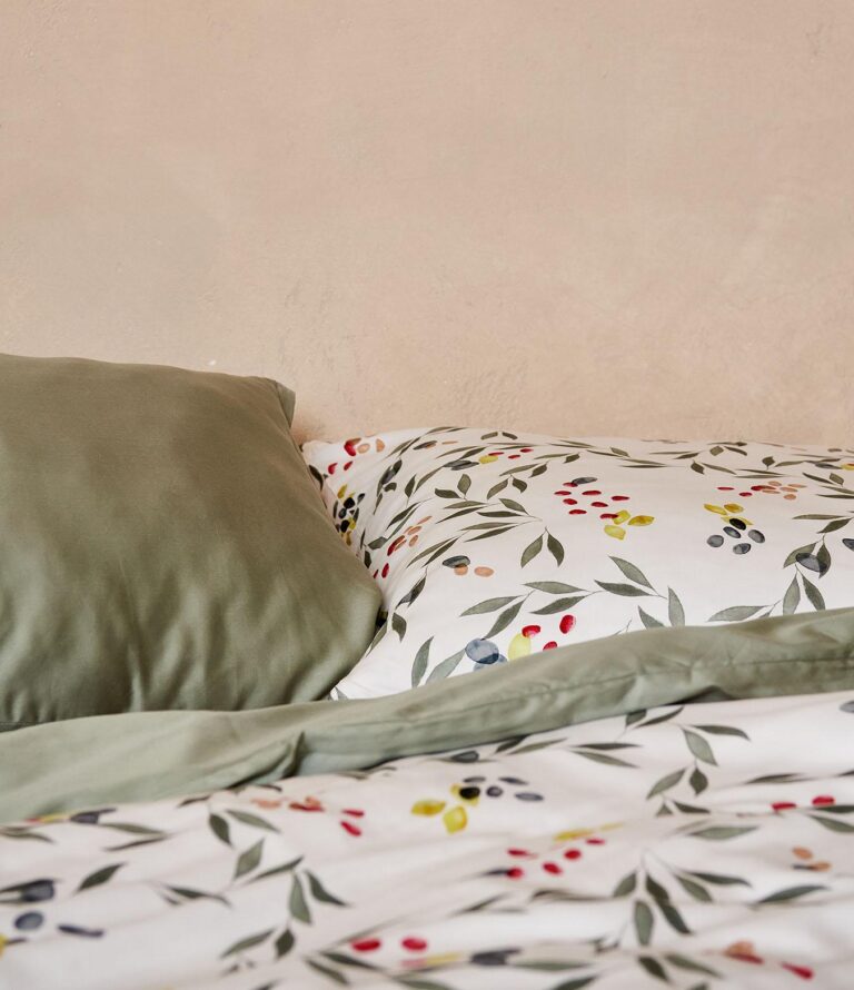 Ein ordentlich gemachtes Bett mit Bettwäsche Journey Living Amiata: ein grüner Kissenbezug und eine Bettdecke, gepaart mit weißen Laken, alles verziert mit einem lebhaften botanischen Blätter- und Beerenmuster, vor einer hellbeigen Wand.