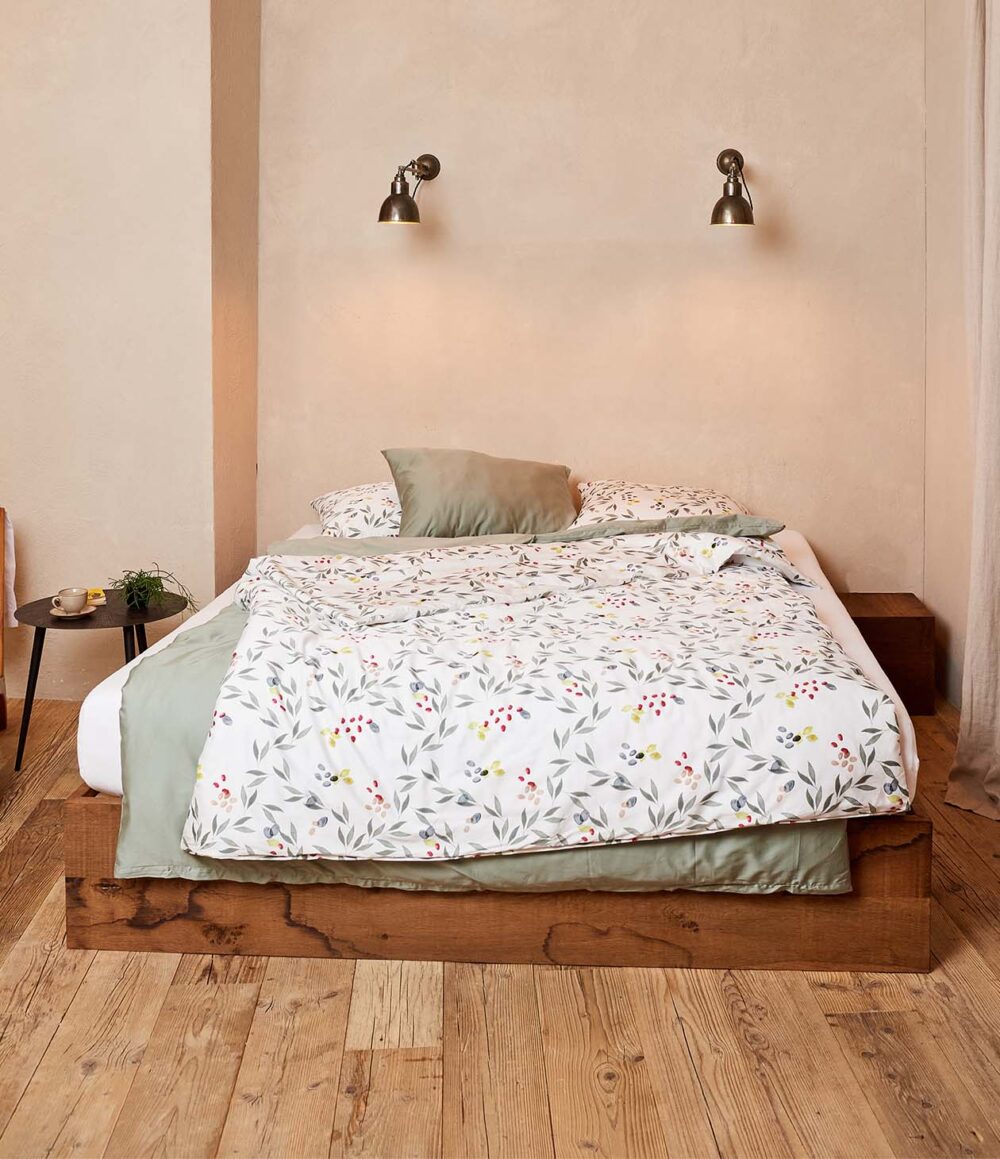 Ein gemütliches Schlafzimmer mit einem Bettgestell aus Holz, Bettwäsche Journey Living Amiata mit Blumen- und Blattmustern, hellgrünen Akzenten, zwei Kissen, Wandleselampen über dem Bett und einem runden Beistelltisch mit einer Pflanze auf Holzfußboden.