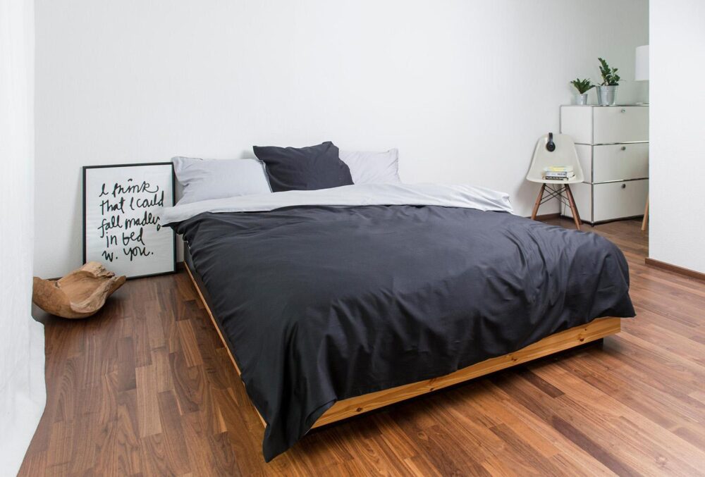 Ein minimalistisches Schlafzimmer mit einem niedrigen Holzbett, Bettwäsche Nobilium Mia in Grau und Weiß, einem gerahmten Zitat, das an der Wand lehnt, einer Topfpflanze, einem Stuhl und einer weißen Kommode auf poliertem Holzboden.