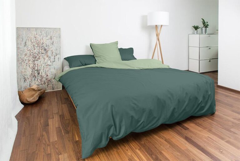 Ein modernes Schlafzimmer mit Holzboden, grüner Bettwäsche Nobilium Mia, zwei Kissen, einer weißen Kommode mit Pflanzen, einer Stehlampe mit weißem Schirm und einem abstrakten Gemälde, das auf dem Boden vor einer weißen Wand steht.