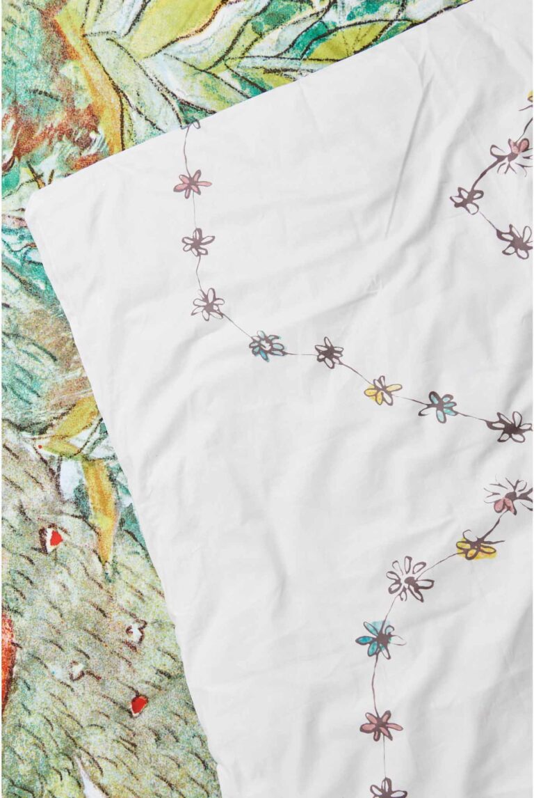 Das weiße Laken Bettwäsche Flurina mit einer Kette aus kleinen bunten Blumen ist teilweise über eine grün-gelbe, blumengemusterte Decke drapiert.